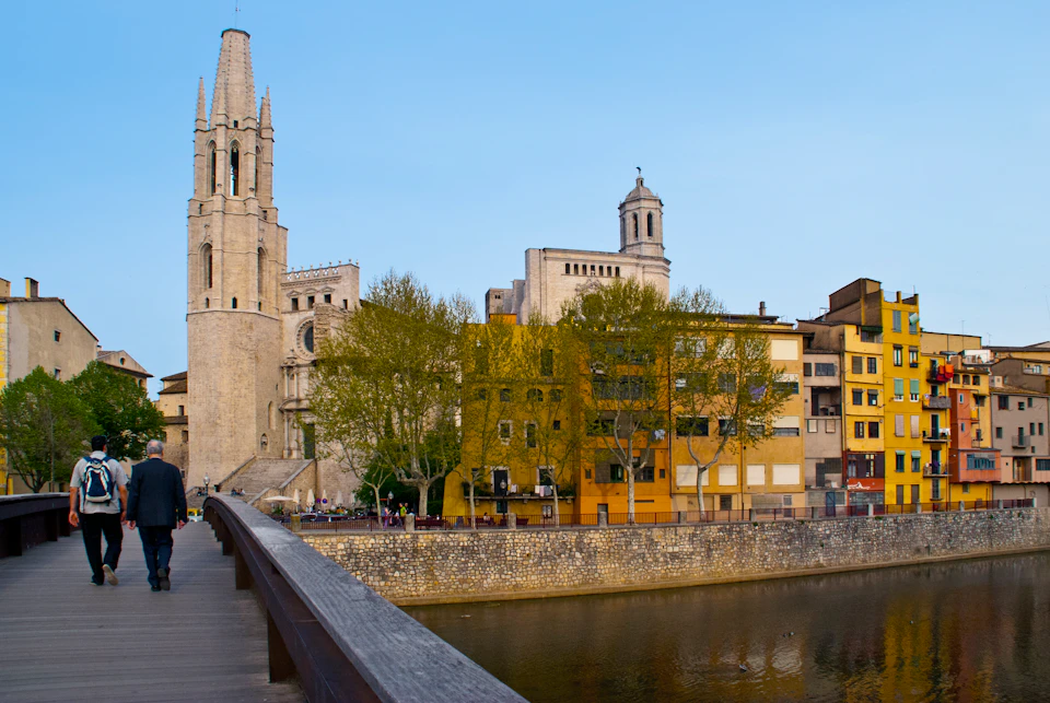 A stone church with a tall bell tower and nearby colorful buildings are visible from a bridge over a river.