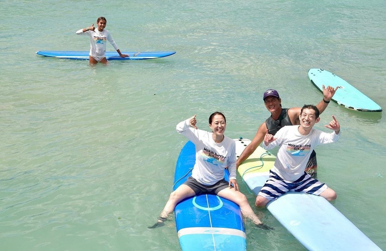 Grupa ludzi w płytkiej wodzie uśmiechających się i pozujących na deskach surfingowych, noszących codzienne ubrania i cieszących się oceanem.