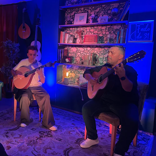 Två musiker spelar gitarrer i ett svagt upplyst rum med en lampa, bokhyllor och en blommig bakgrund medan två kvinnor tittar på och håller glasögon.