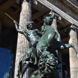 Bronsstaty av en man på en uppfödningshäst med ett spjut, framför en historisk byggnad med stora kolonner och latinsk inskription.