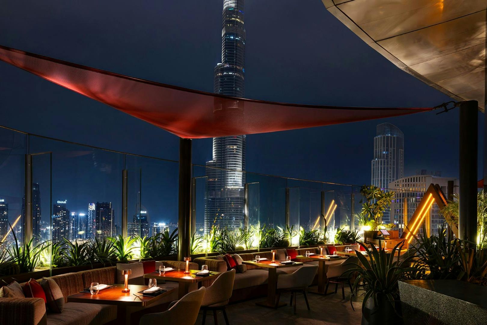 Rooftop restaurant at night with candlelit tables and red canopy, overlooking a city skyline with a prominent tall building.