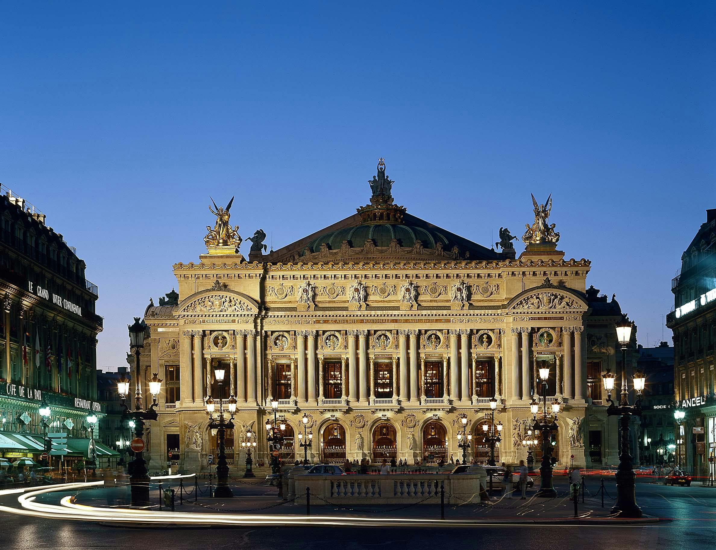 Elaborate, illuminated facade of an ornate building with statues, arches, and columns under a twilight sky.