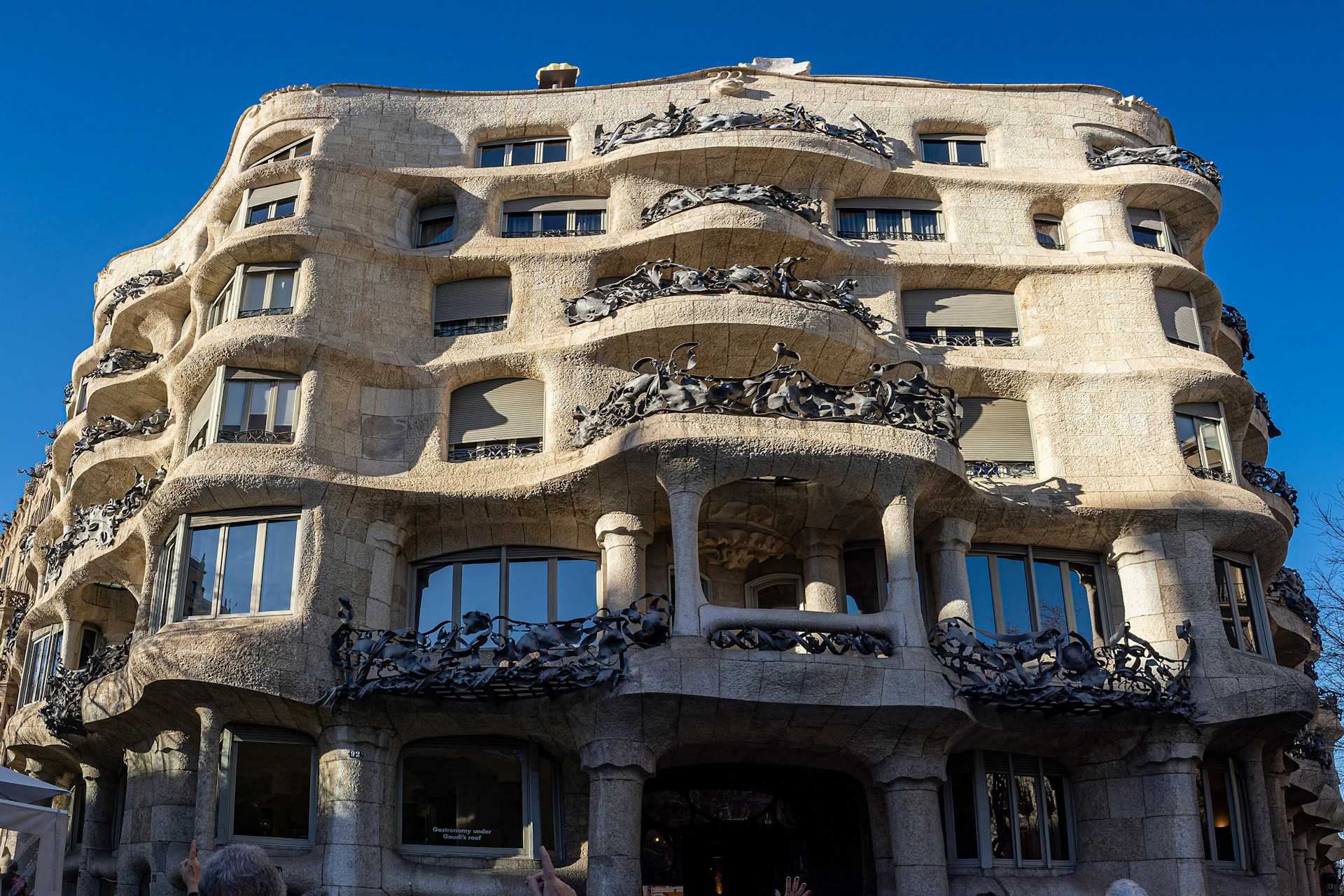 A wavy stone facade of a building with ornate ironwork balconies, viewed under a clear blue sky.