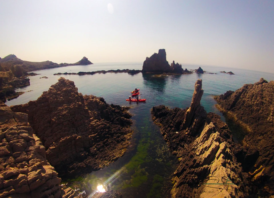 People kayaking amongst rocky formations on a calm sea beneath a clear sky. Rugged cliffs frame the scene.