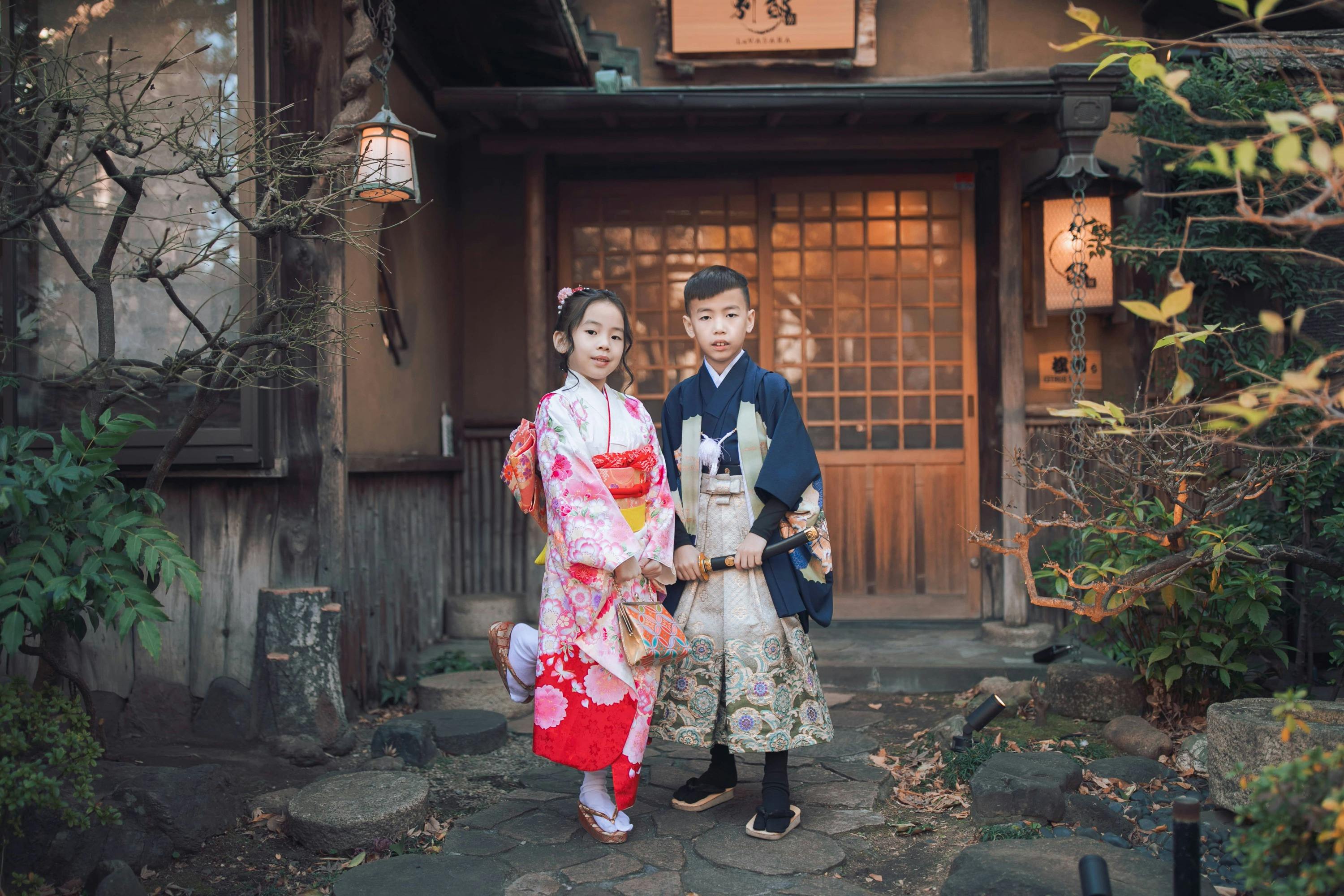 Two children in traditional Japanese attire stand in front of a wooden building with a sliding door and lanterns.