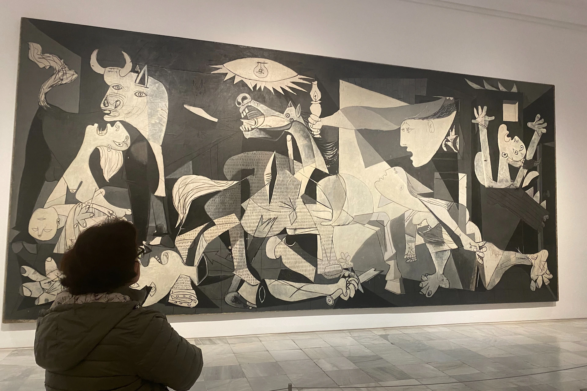 A person observes Picasso's painting 'Guernica' displayed on a museum wall, featuring cubist depictions of figures and animals.