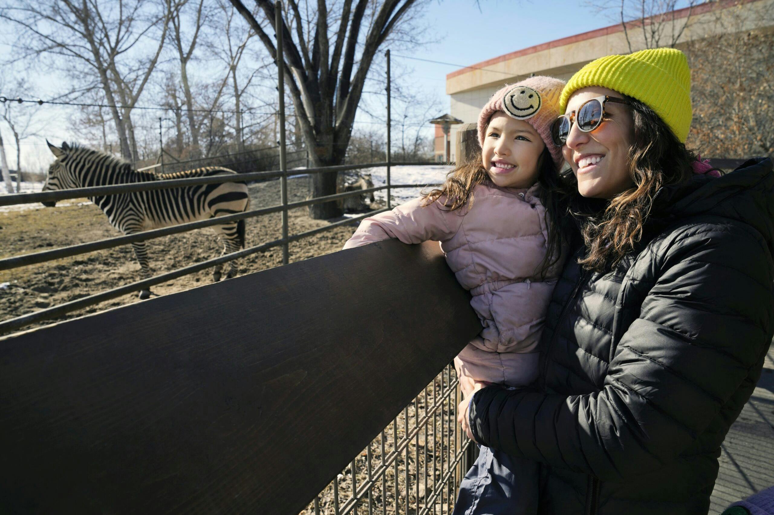 모자와 재킷을 포함한 겨울 옷을 입은 미소 짓는 여성과 어린이가 화창한 날 야외 울타리 옆에 서 있습니다.