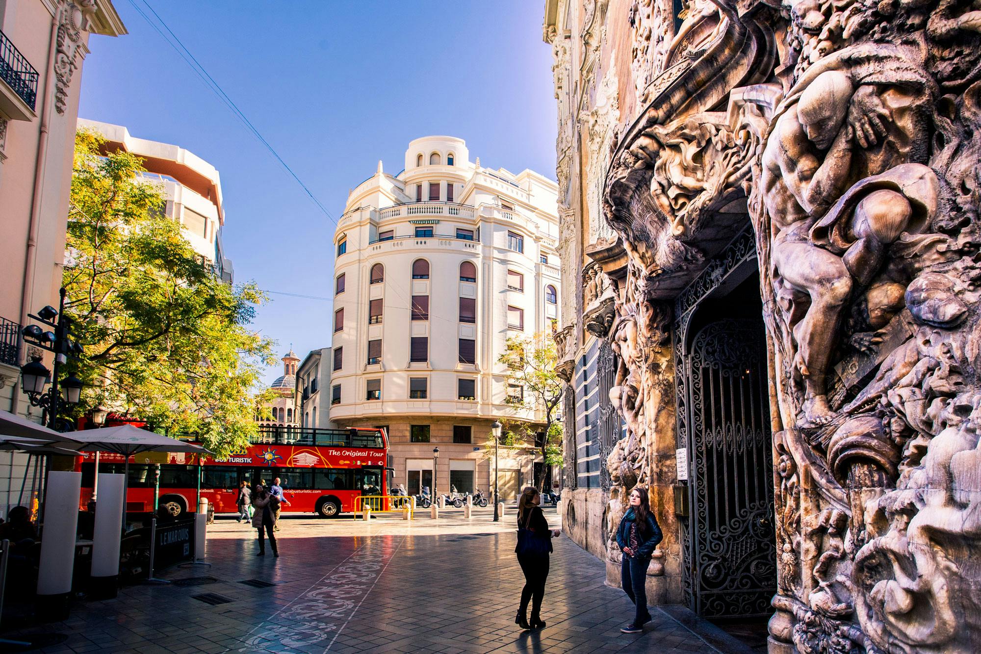 Urban street with ornate building facade, red double-decker tour bus, people walking, and a rounded corner building in the background.