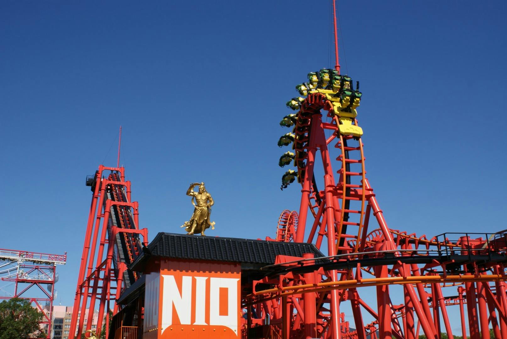 A red rollercoaster with loops and a golden statue on a building marked 'NIO' under a clear blue sky.