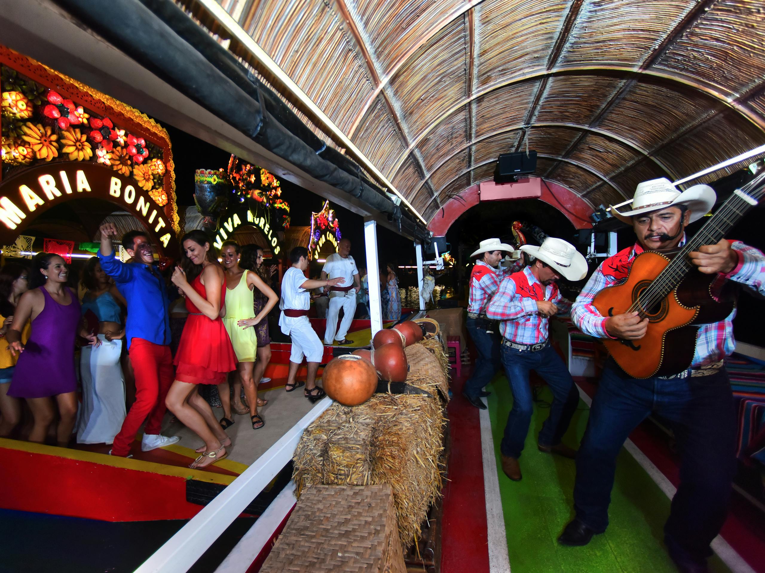 Μουσικοί με καουμπόικα καπέλα παίζουν κιθάρες σε βάρκα κάτω από αχυρένια στέγη, ενώ άνθρωποι με πολύχρωμα ενδύματα χορεύουν και απολαμβάνουν την ατμόσφαιρα.