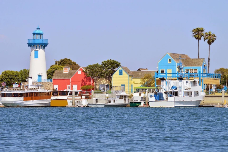 Colorful buildings and boats line a waterfront, with a lighthouse on the left and a clear blue sky above.
