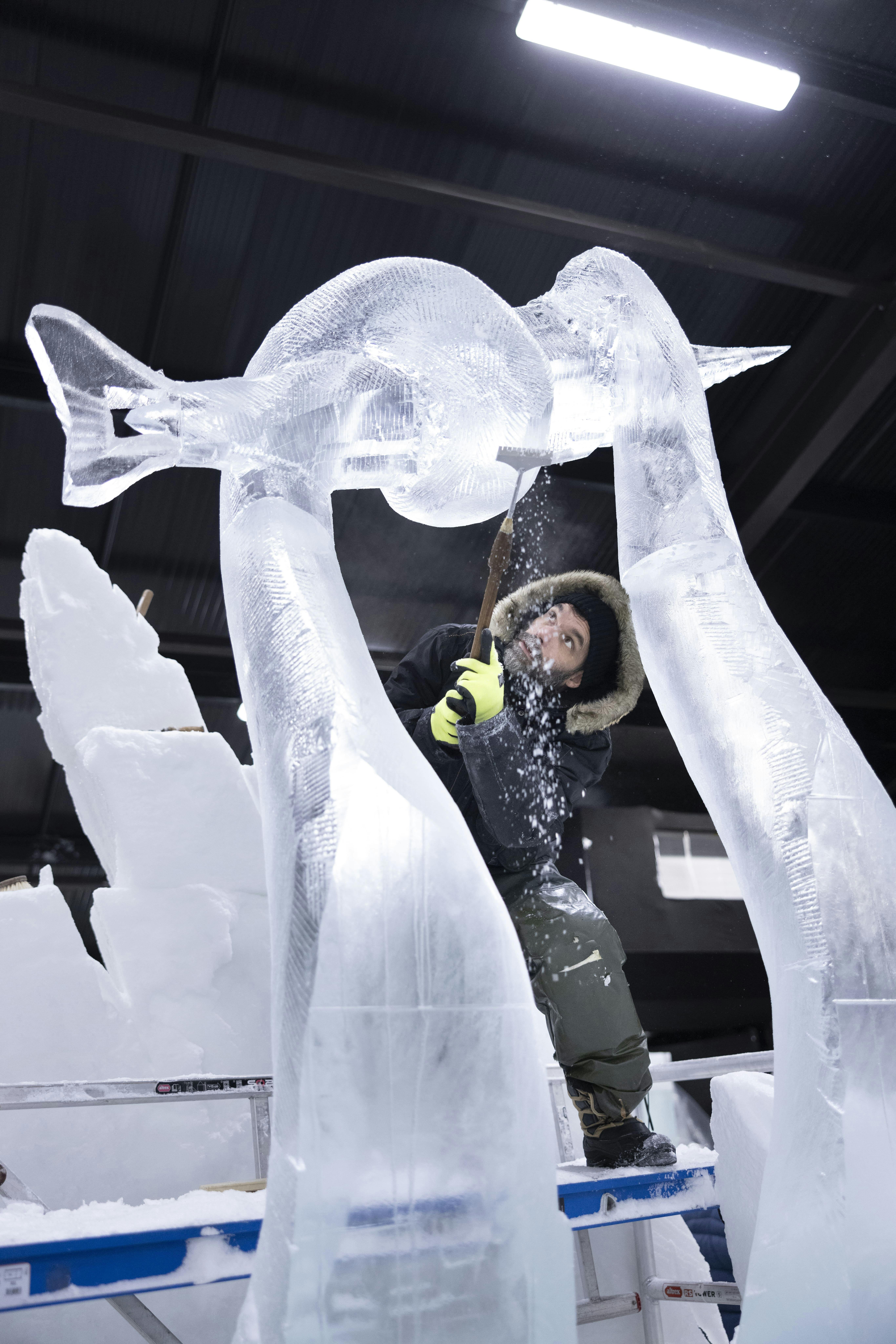 Beleuchtete Eisskulpturen einer Person, die sich einem geflügelten Wesen mit menschlichem Gesicht, einer Krone und detaillierten Flügeln zuwendet.