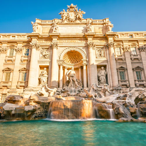 Fontän i barockstil med invecklade skulpturer och kaskadvatten framför en stor, utsmyckad byggnad under en klarblå himmel.