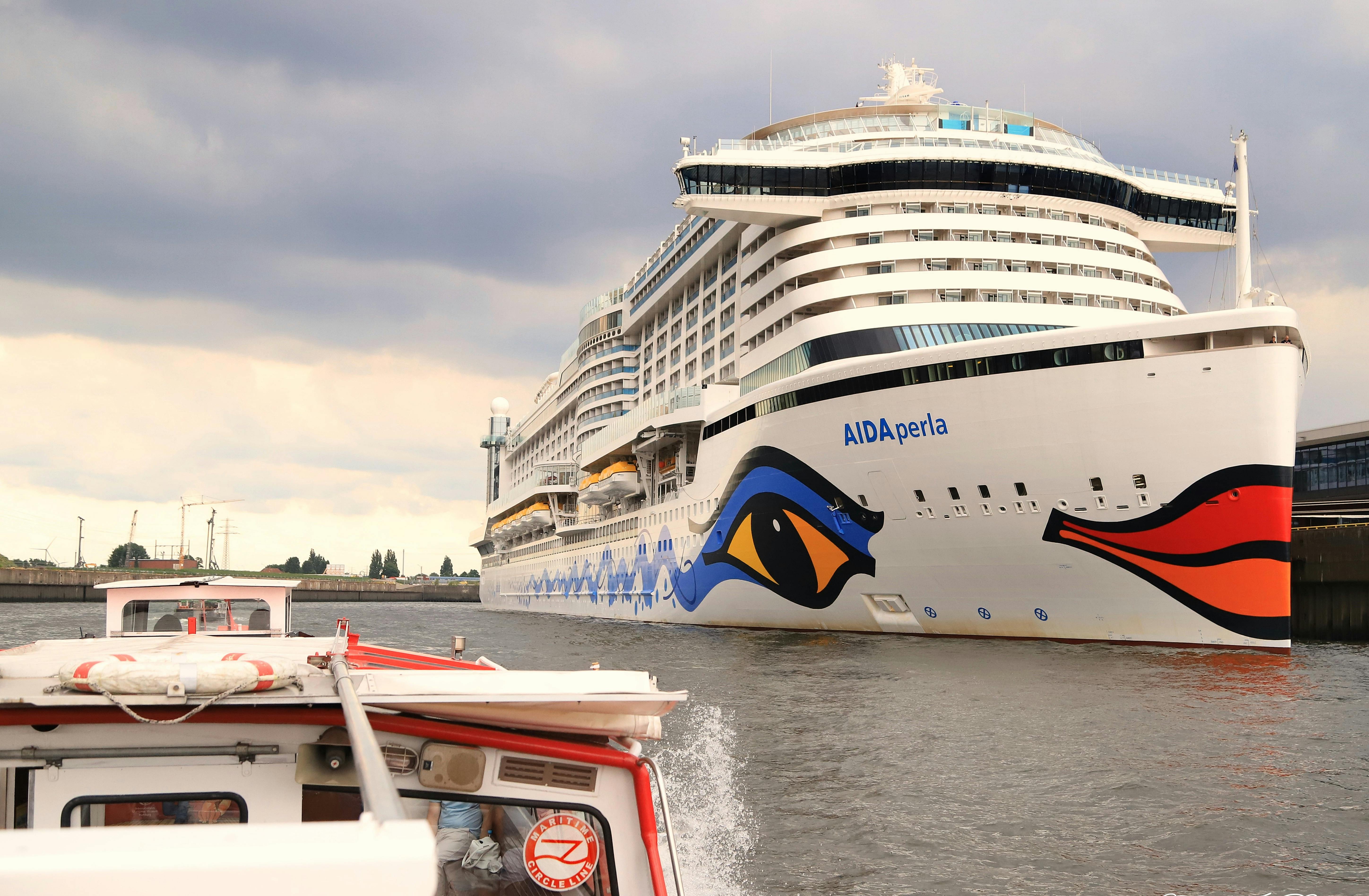 Grand bateau de croisière nommé "AIDAperla" naviguant sur une rivière à côté d'un plus petit bateau à moteur par une journée nuageuse.