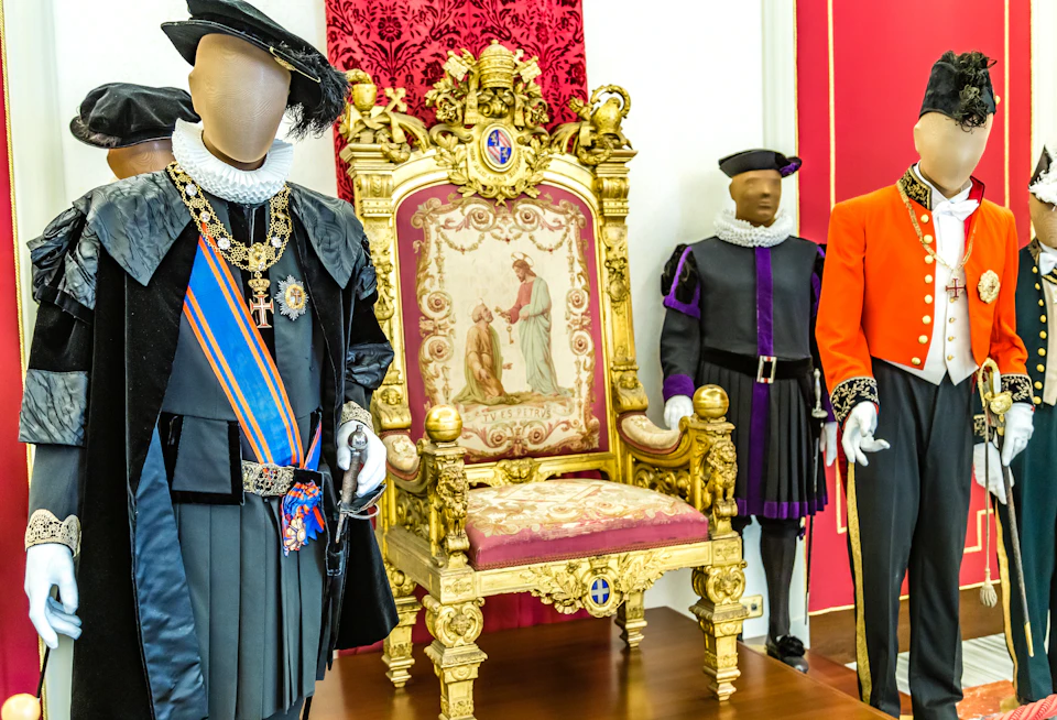 A display of ornate costumes, including a golden throne, a black robe, a black outfit with a purple sash, and a red uniform with medals.