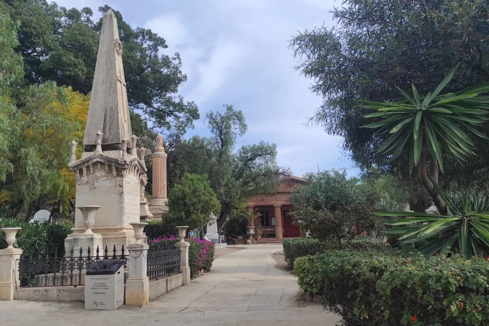 Outdoor park scene with a stone pathway surrounded by trees, bushes, and monuments. A terracotta building is visible in the background.