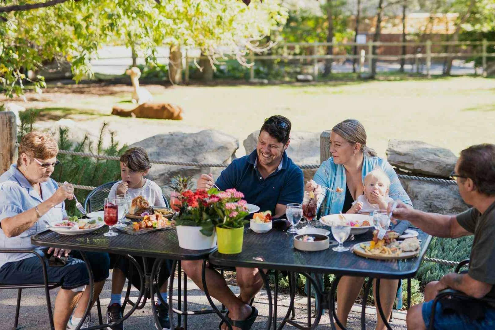 아기를 포함한 4명이 야외 테이블에 둘러앉아 식사를 즐기고 있습니다. 테이블에는 음식, 음료, 꽃이 준비되어 있습니다.