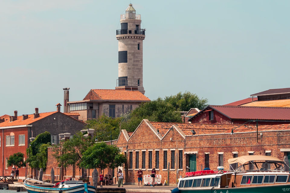 A lighthouse stands behind brick buildings by a waterfront, with trees and boats. Several people are visible near the buildings.