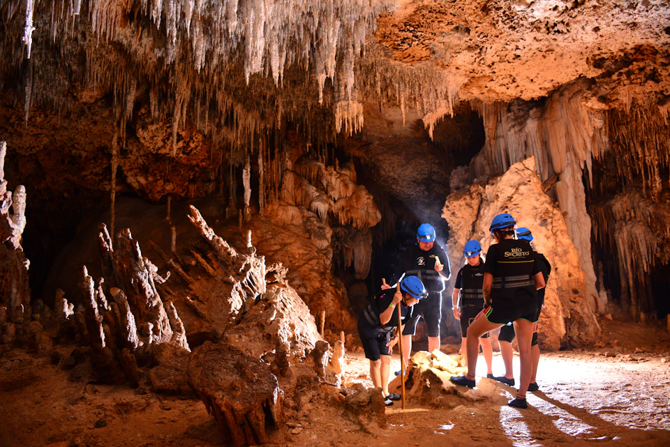 Four people in helmets explore a dimly lit cave with stalactites and stalagmites, examining formations on the ground.