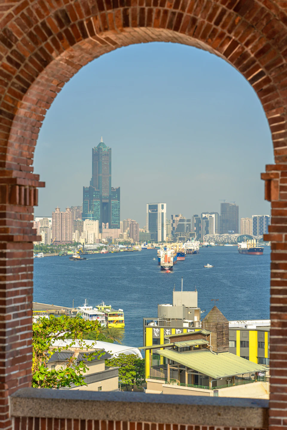A cityscape view through a brick archway, showcasing a river, boats, and tall buildings under a clear sky.