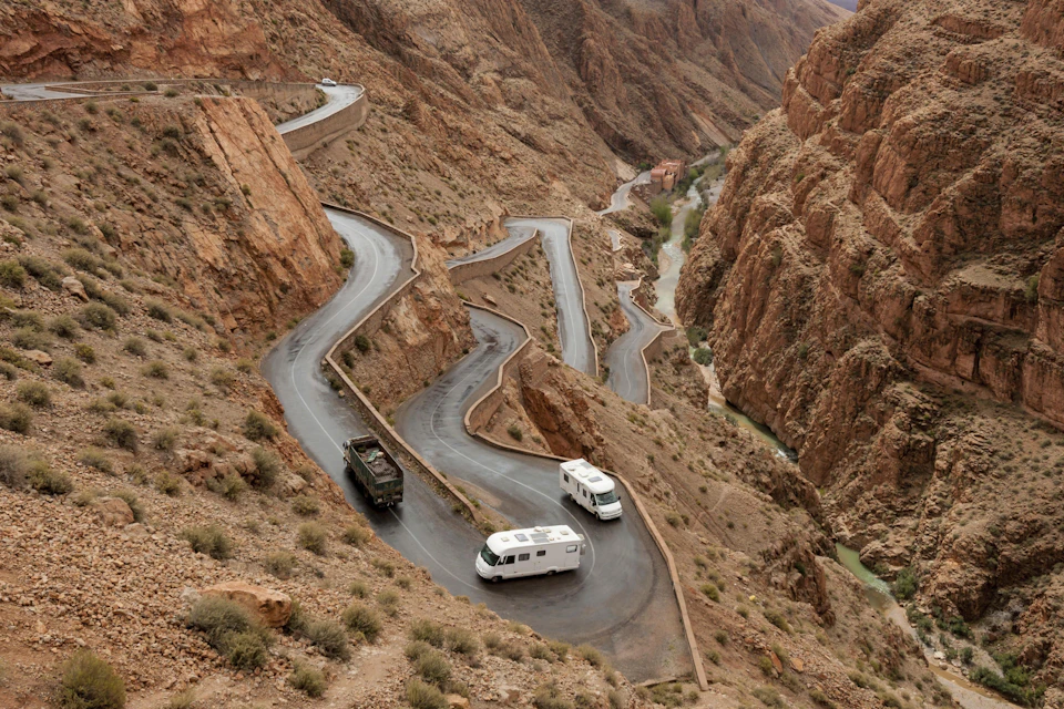 Winding mountain road with sharp curves, two white RVs, and a dark vehicle, surrounded by rocky terrain and a narrow river below.