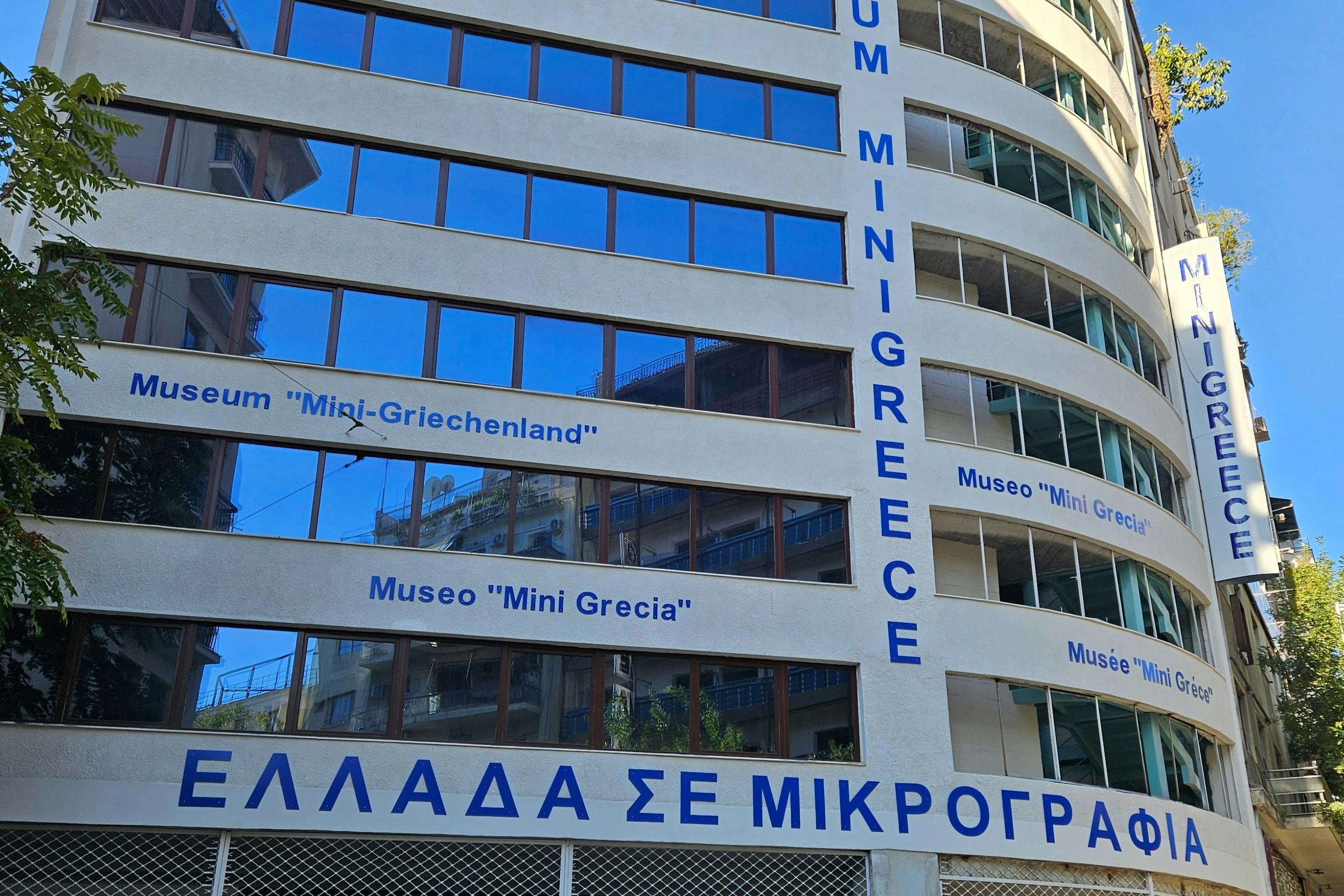 Facade of a building with blue-framed windows and multilingual signs for "Museo Mini Grecia." Text in Greek and other languages is visible.