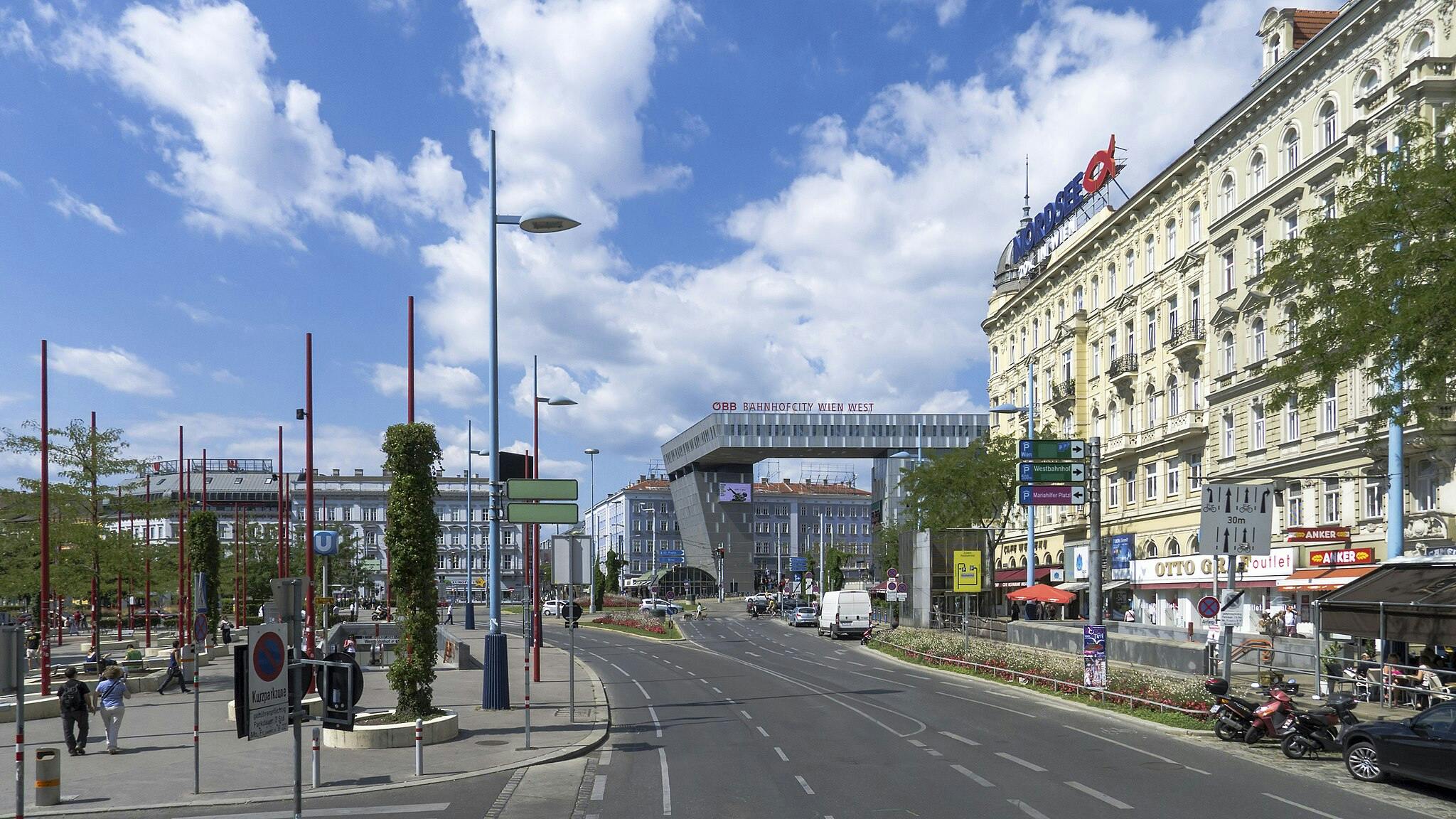 Veduta stradale di una vivace area cittadina con un edificio moderno con la scritta "Bahnhofcity Wien West", negozi, pedoni e alcune motociclette parcheggiate.
