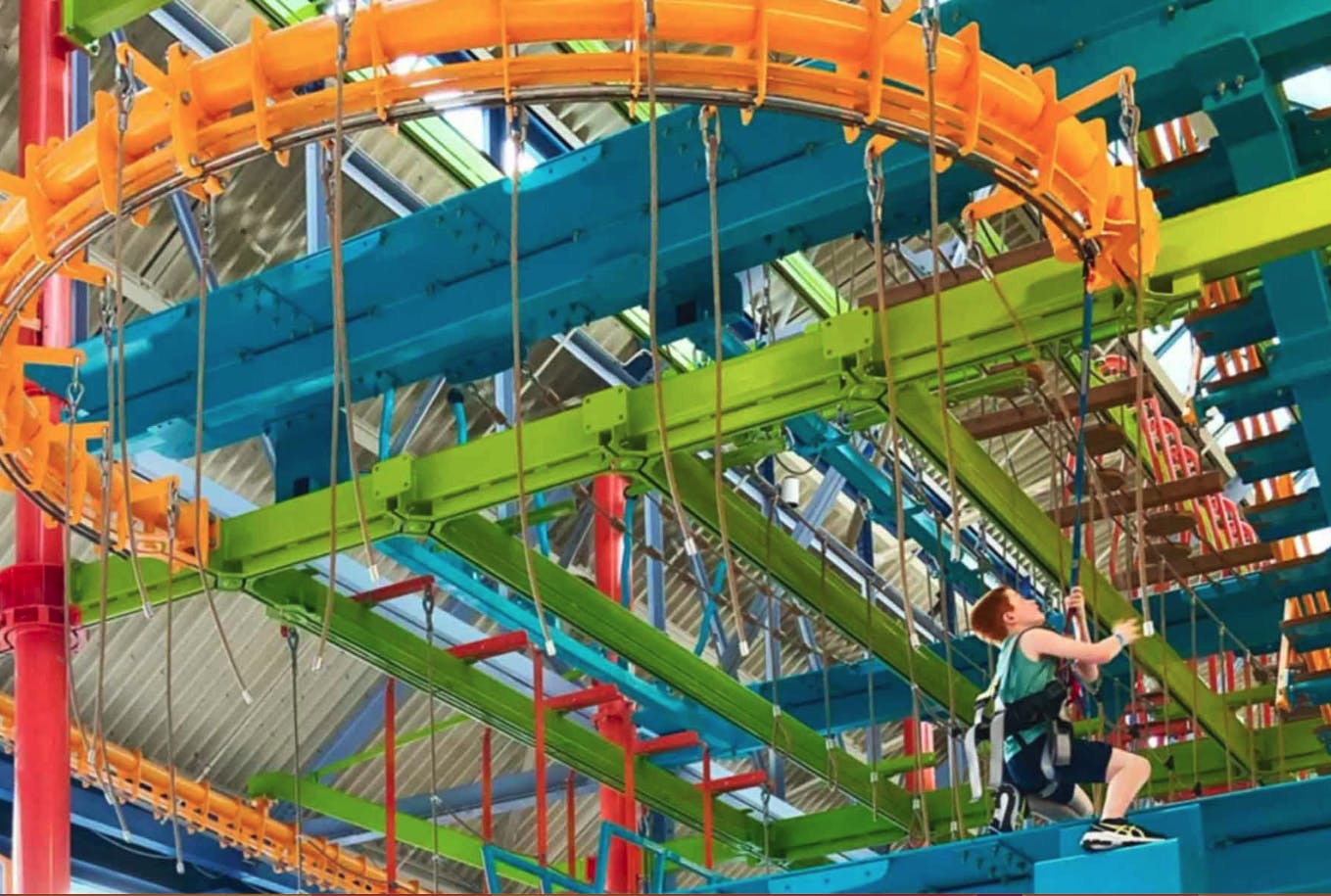 한 어린 소년이 파란색, 녹색, 주황색, 빨간색 구조물과 안전 장치가 있는 다채로운 실내 로프 코스를 올라갑니다.