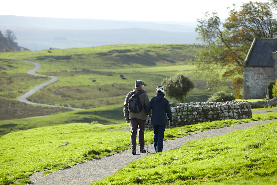 Two people with walking sticks hike a paved path through a green, hilly landscape on a sunny day.