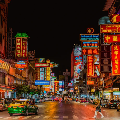 Belebte nächtliche Straße in einem Chinatown, gesäumt von bunten Leuchtreklamen in verschiedenen Sprachen, Autos und kreuzenden Fußgängern.