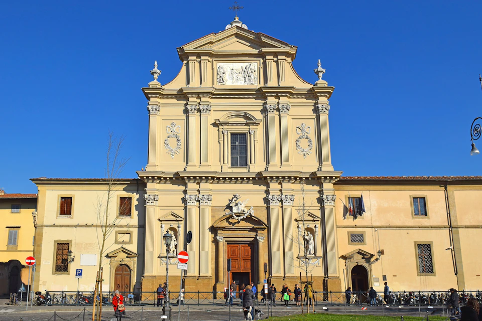 Ornate church facade with statues and intricate details under a clear blue sky. People walk and gather near the entrance.