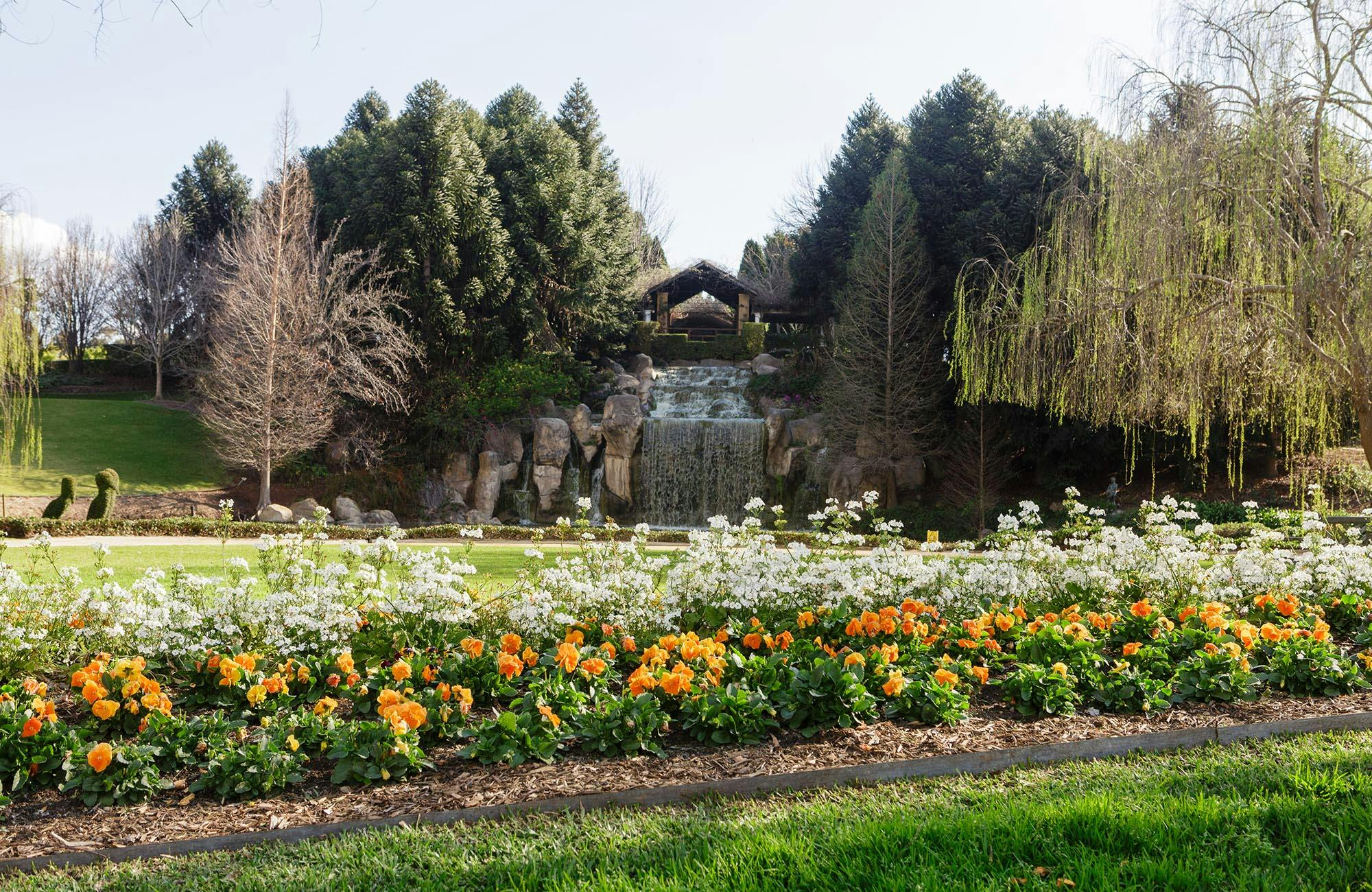 A landscaped garden with colorful flowers in the foreground, a waterfall in the center, and trees in the background.