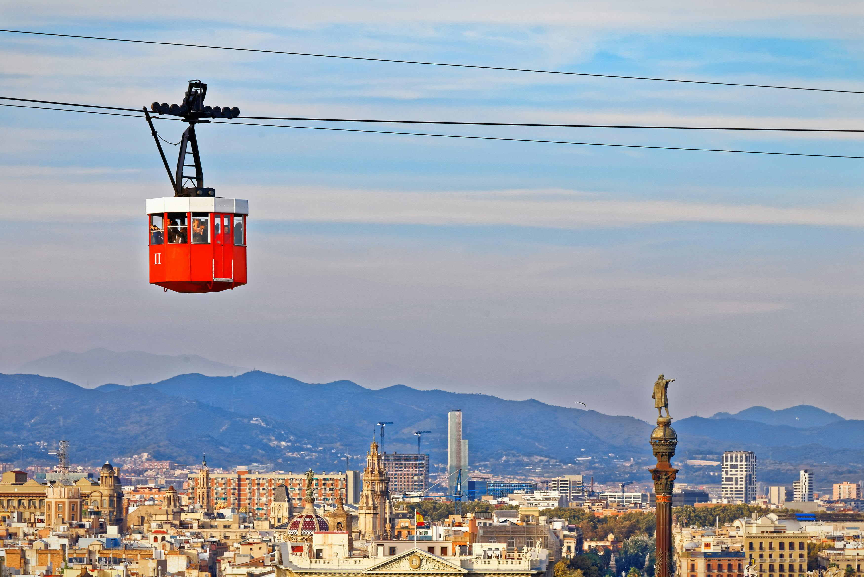 Funivia rossa con passeggeri sospesi su un paesaggio urbano, montagne e un monumento con una statua in un cielo blu.