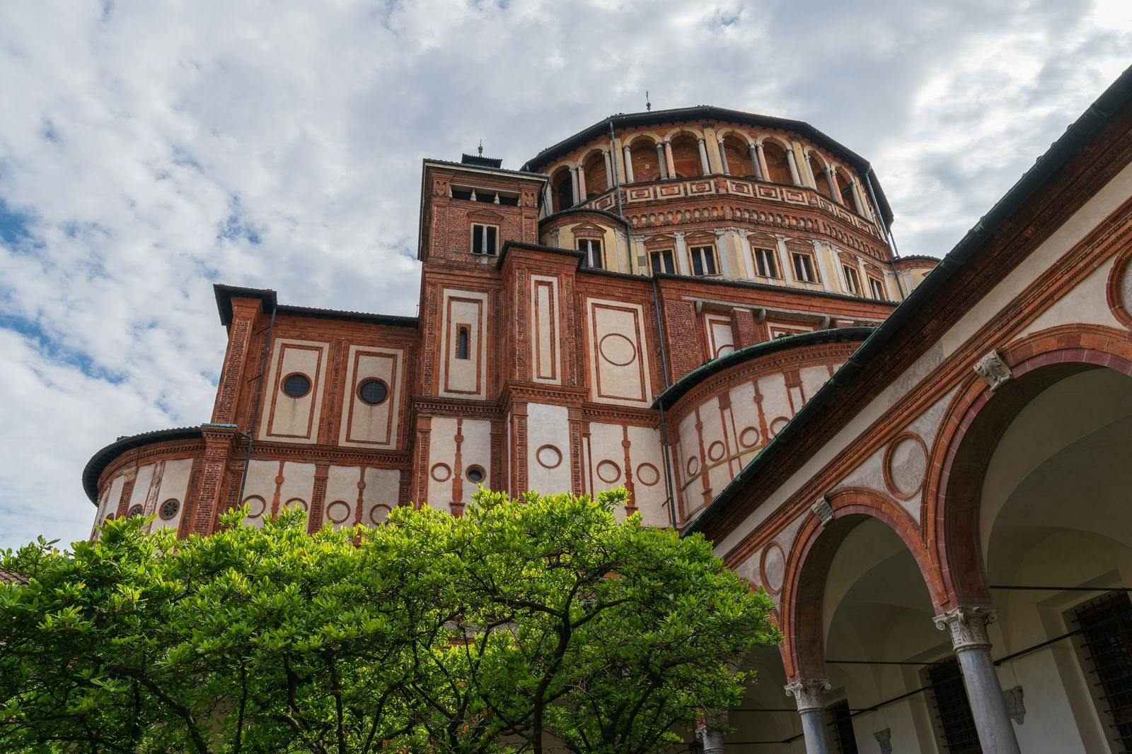 A historic brick building with intricate patterns and an arched roof, surrounded by greenery under a cloudy sky.
