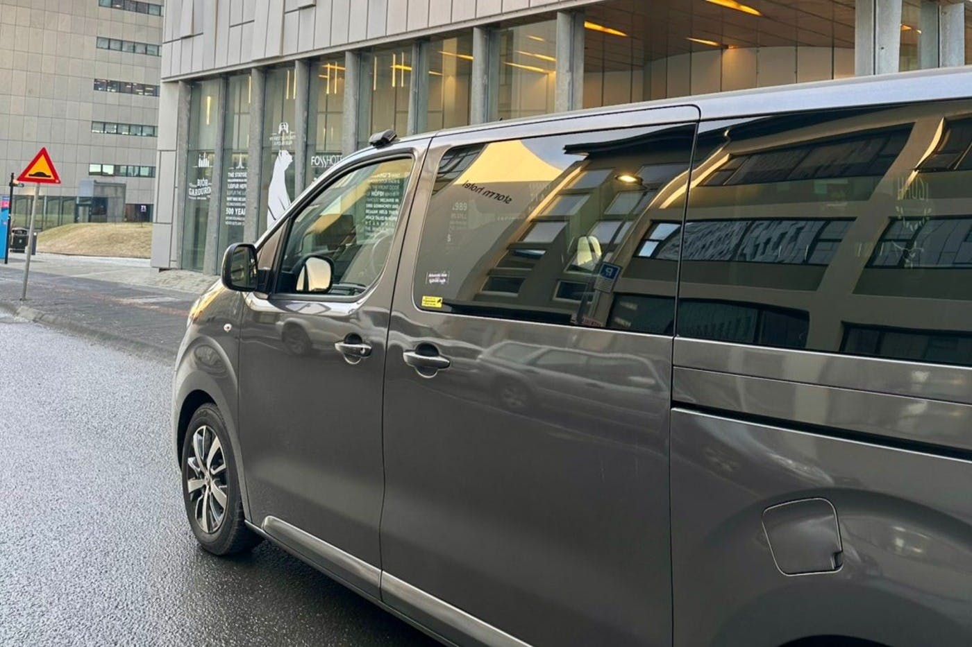 A gray minivan is parked on a street in front of a modern building with large windows and lit interior.