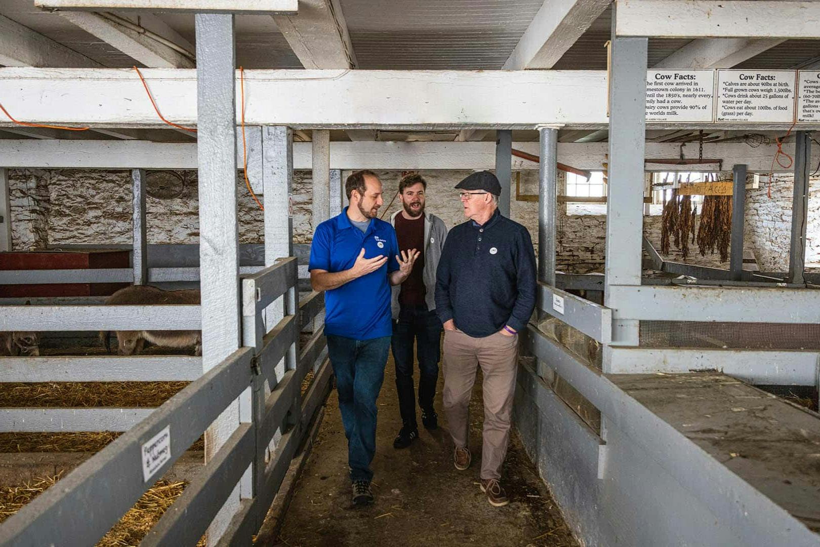 Three men walking and talking in a barn-like building with gray wooden railings and visible stalls.