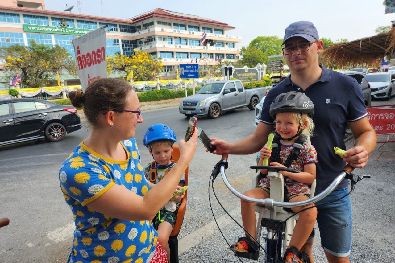 Uma família com duas crianças, uma delas numa cadeira de criança para bicicletas, ao ar livre perto de uma estrada com um edifício ao fundo.