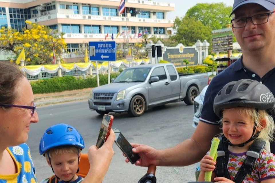 A family with two children, one in a child bike seat, outdoors near a road with a building in the background.