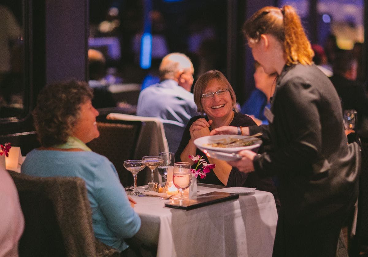Two women smiling at a restaurant table while a server delivers a dish. The table has wine glasses, candles, and flowers.