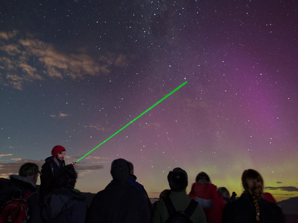 A group of people stargazing, with one person pointing a green laser towards the night sky illuminated by stars and auroras.
