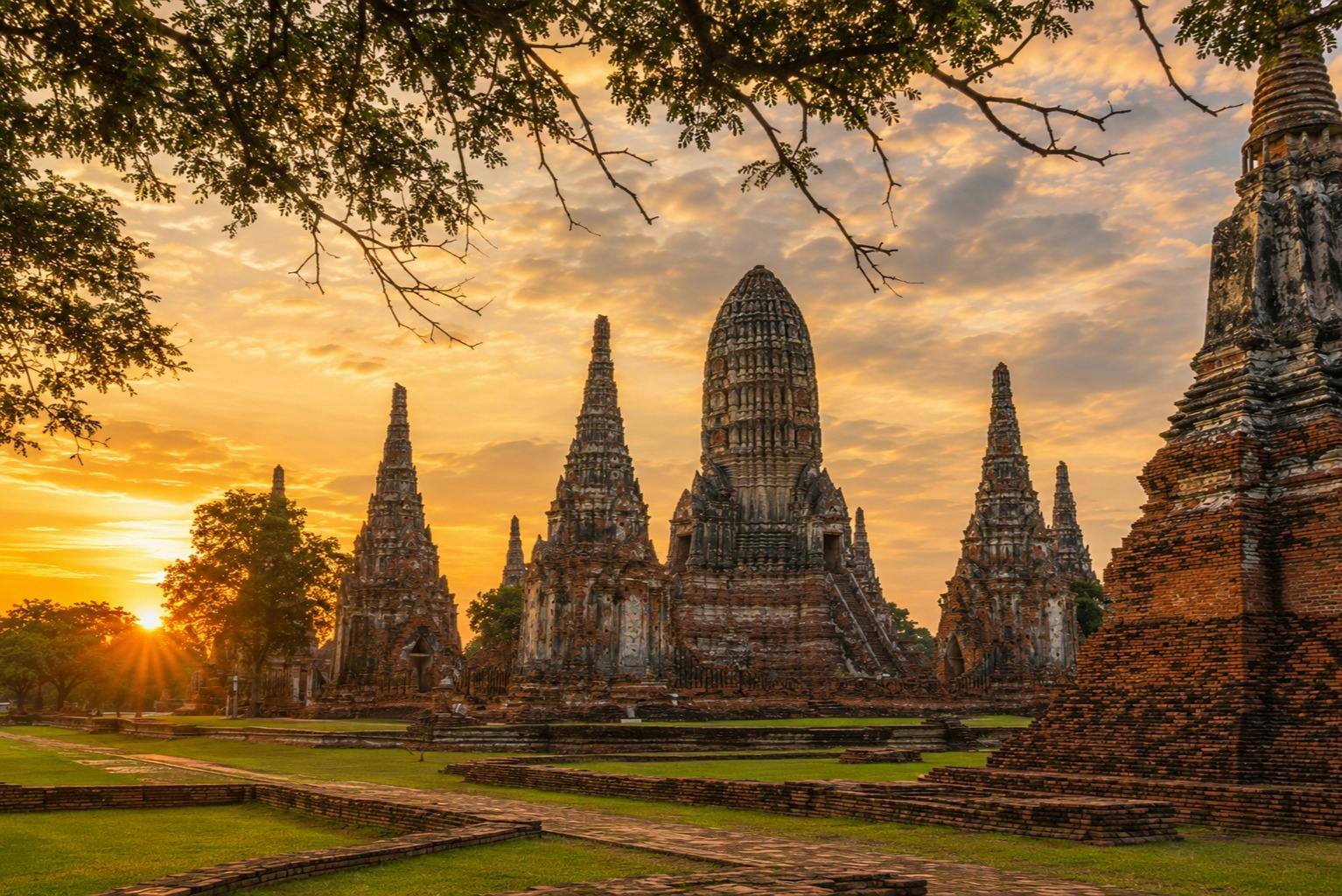 Ancient temple ruins with tall spires at sunset, surrounded by green grass and framed by tree branches.