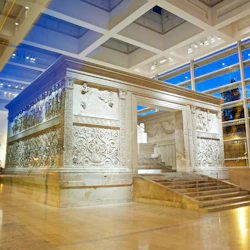 En detaljerad marmorstruktur med sniderier, innesluten i en glasbyggnad med högt i tak och stora fönster som avslöjar en blå himmel.