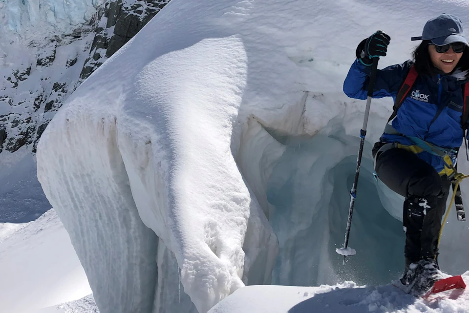 A person in hiking gear is standing on snow near an ice formation, holding a pole, and smiling under a sunny sky.