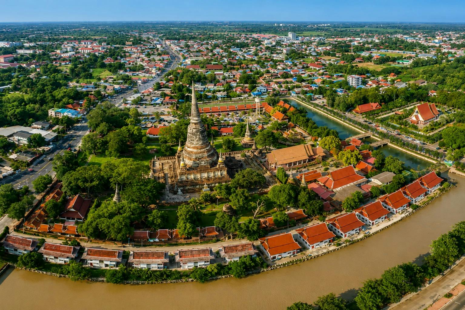Aerial view of a temple complex surrounded by trees, a cityscape, and a river in a sunny area.