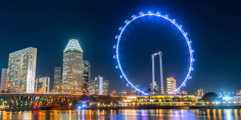 Horizonte da cidade à noite com arranha-céus iluminados e uma grande roda gigante perto de uma zona ribeirinha que reflecte luzes coloridas.