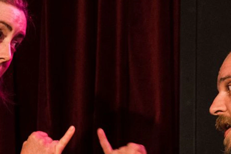 Two people perform on stage; one stands gesturing while the other sits, both engaged in animated interaction.