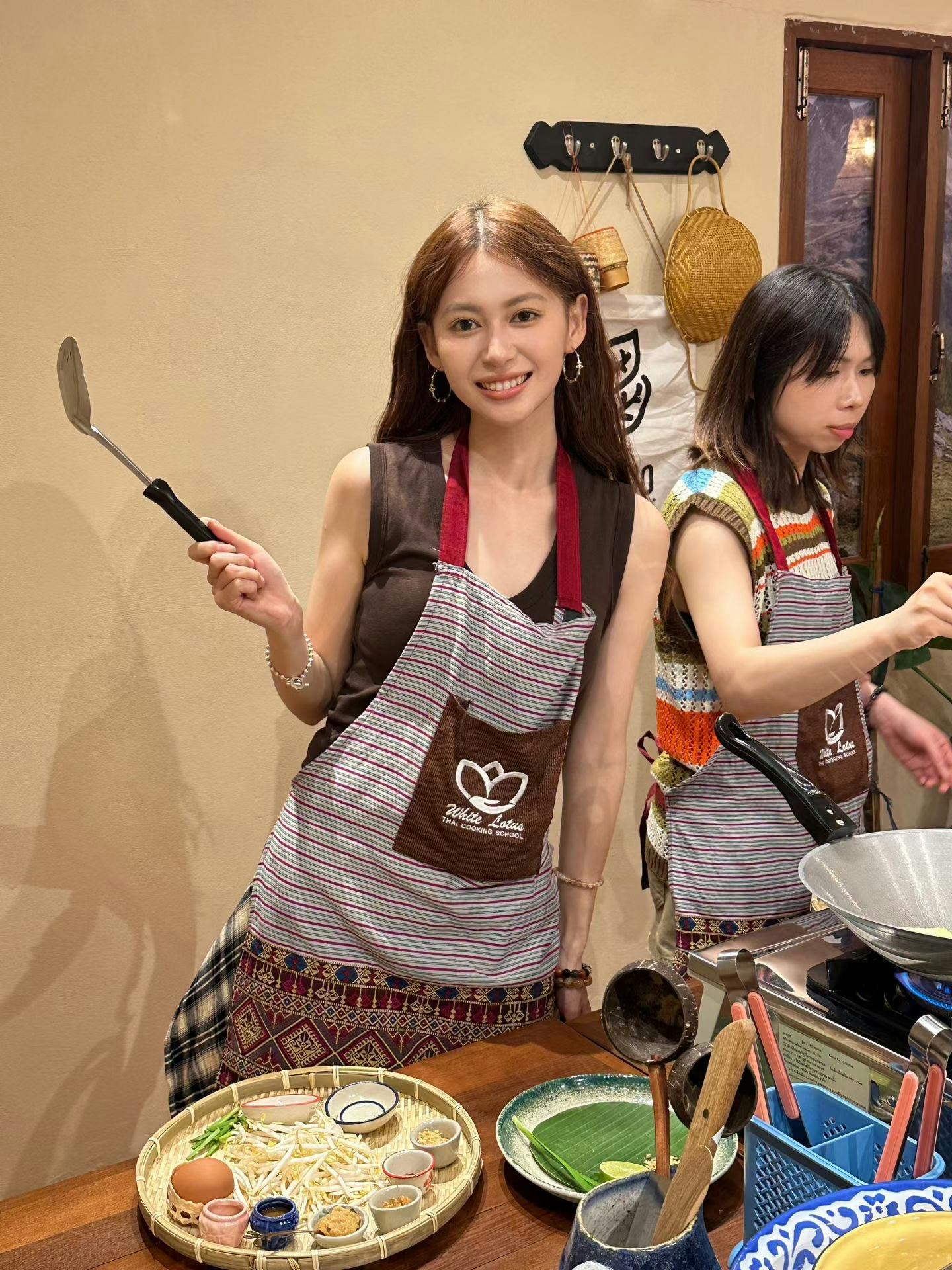 Two women in aprons cooking in a kitchen, one holding a spatula and smiling at the camera.