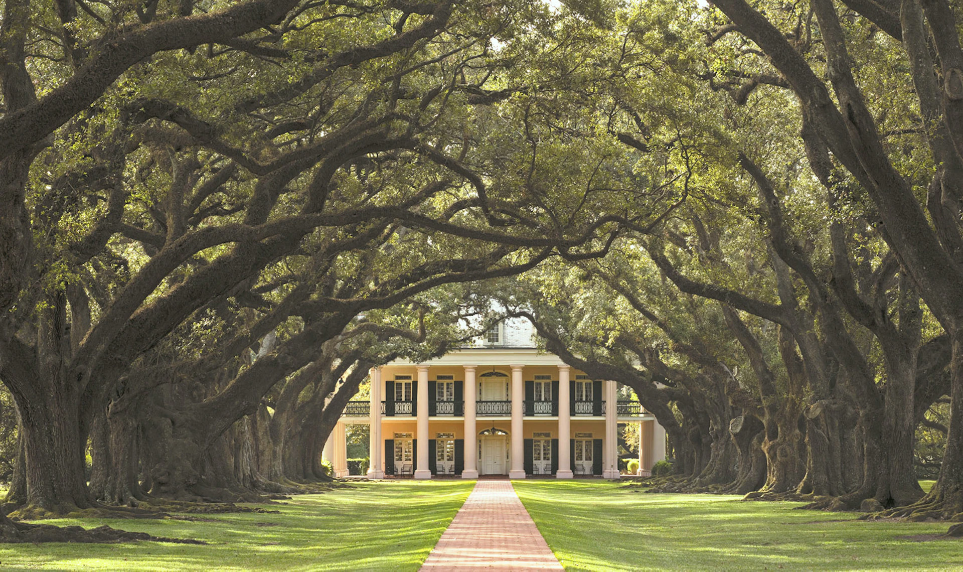 Large, symmetrical two-story house with columns, framed by arching oak trees and a red brick pathway leading to the entrance.