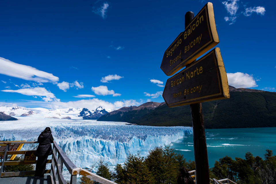 A glacier with a turquoise lake, snowy mountains, and a wooden sign marking viewing balconies under a blue sky.