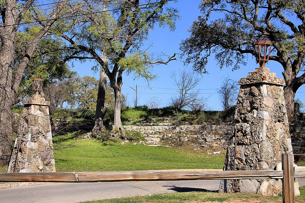 Dos pilars de pedra amb llanternes flanquegen una tanca de fusta en un parc amb arbres i un cel blau clar.